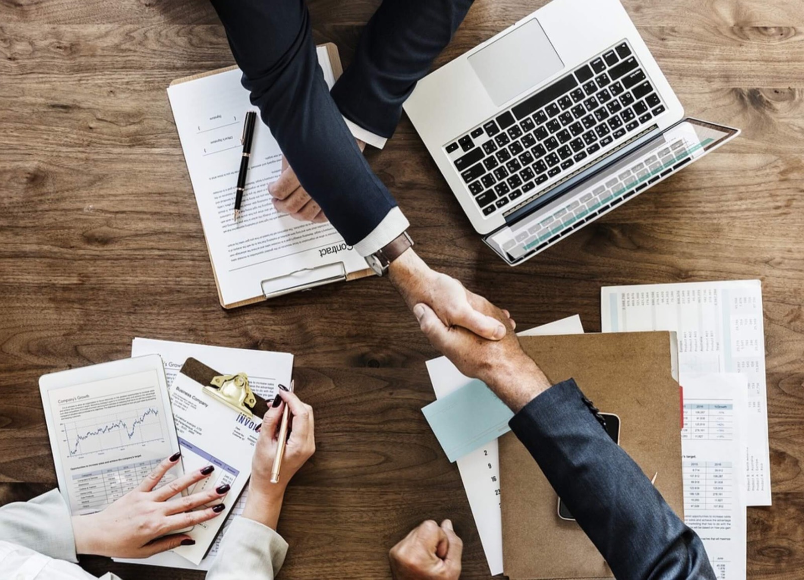 Due professionisti si stringono la mano su una scrivania con documenti e un portatile